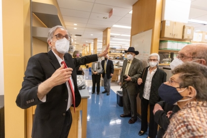 Standing in a laboratory space, a person wearing a black suit and red tie and face mask gestures while speaking to a crowd of onlookers.